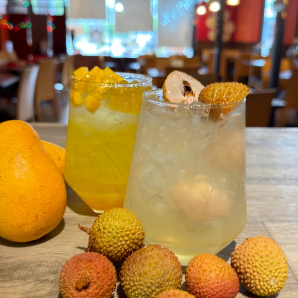 Two refreshing drinks, one mango and one lychee, with fresh fruit on a wooden table.