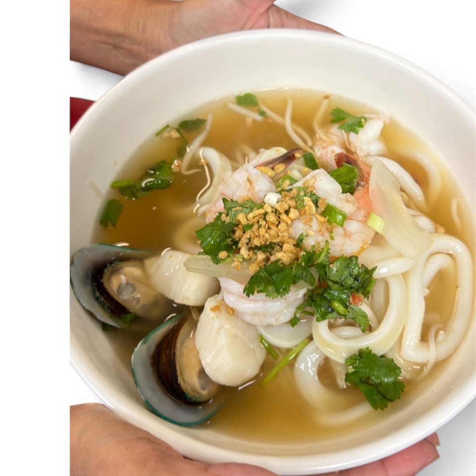 A white bowl of seafood noodle soup with shrimp, mussels, squid, and scallops, garnished with herbs.