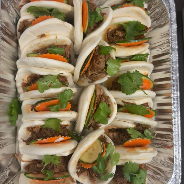 Rows of bao buns filled with shredded meat, cucumber, carrots, and cilantro in a foil tray.