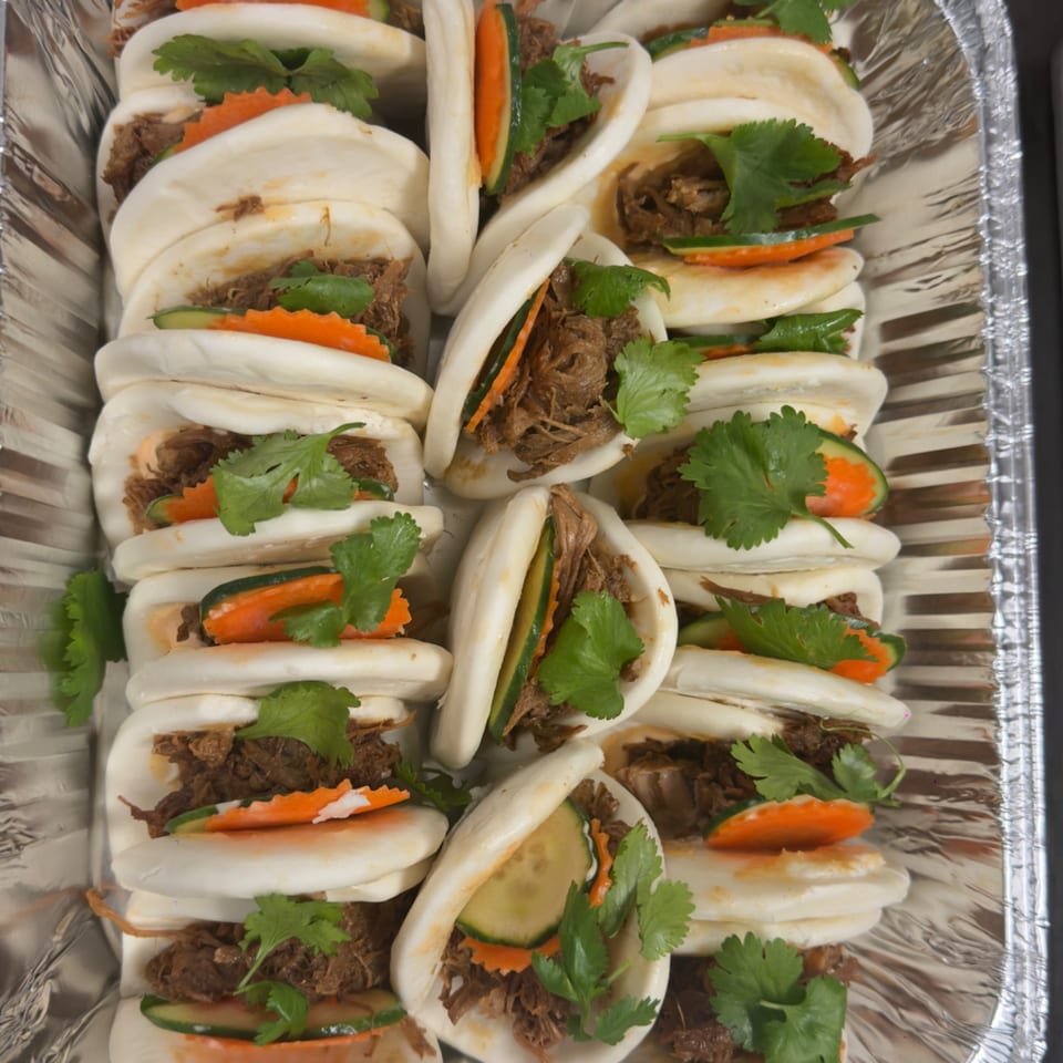 Rows of bao buns filled with shredded meat, cucumber, carrots, and cilantro in a foil tray.